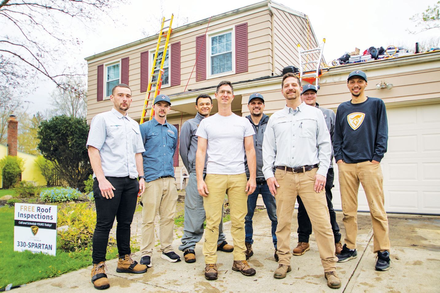 A group of eight people in work attire stand in front of a residential house with a ladder and roofing supplies visible.