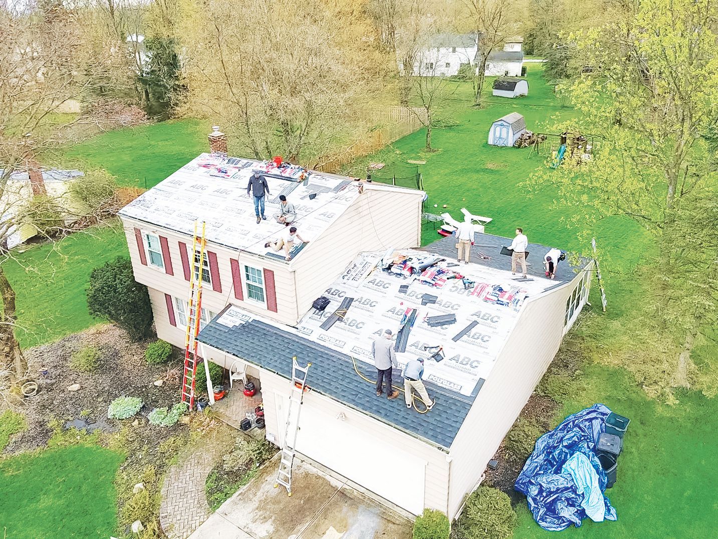 Aerial view of a crew of workers installing new shingles on a suburban house roof surrounded by trees and a green lawn.