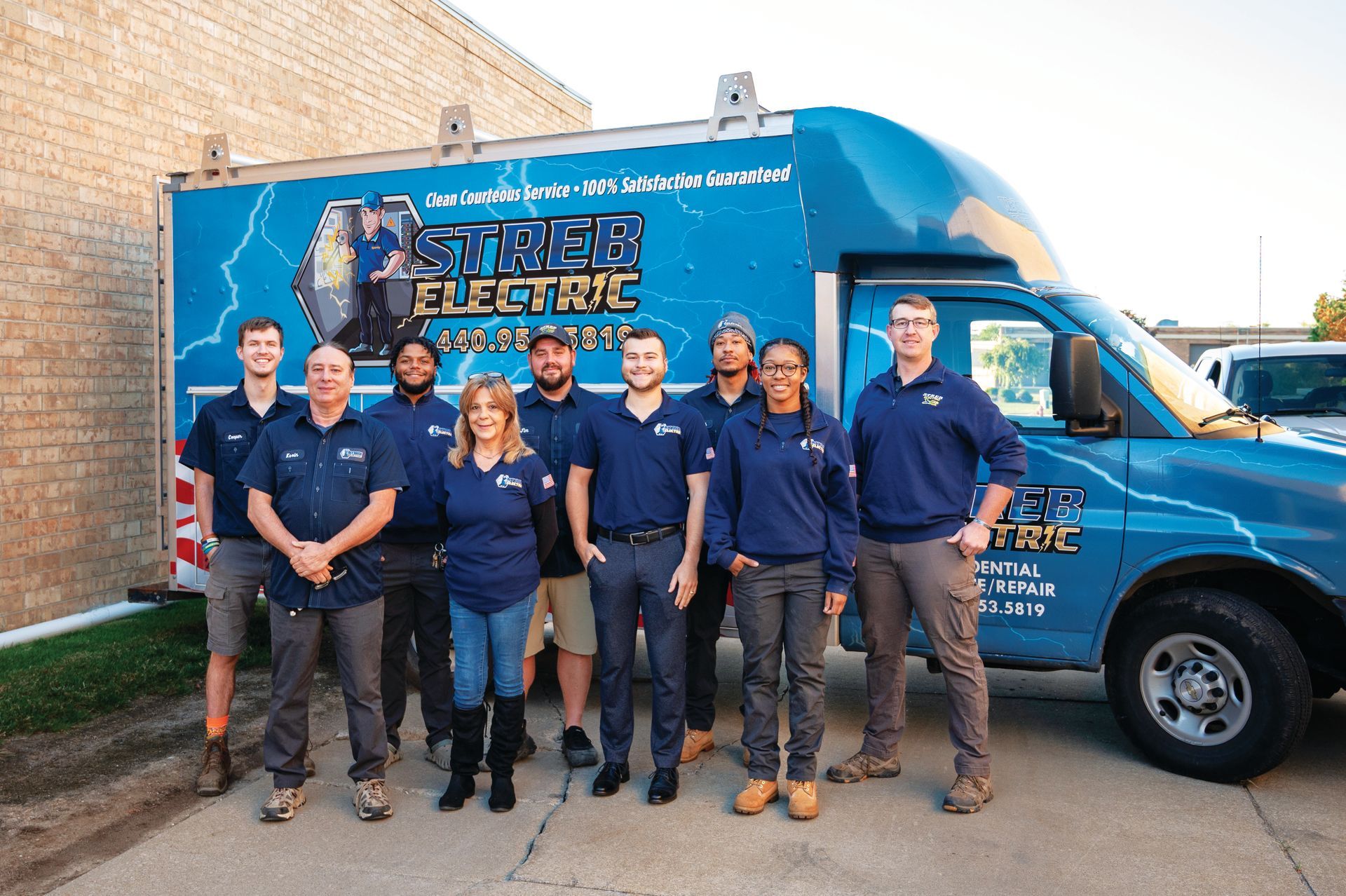 A group of employees in navy blue uniforms stands in front of a blue Sage Electric service truck.