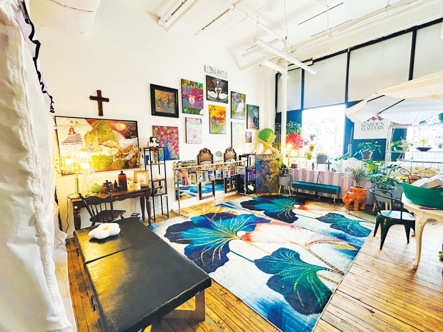 A bright, eclectic room features a large, colorful floral rug, art-covered walls, and a black desk on a wooden floor.