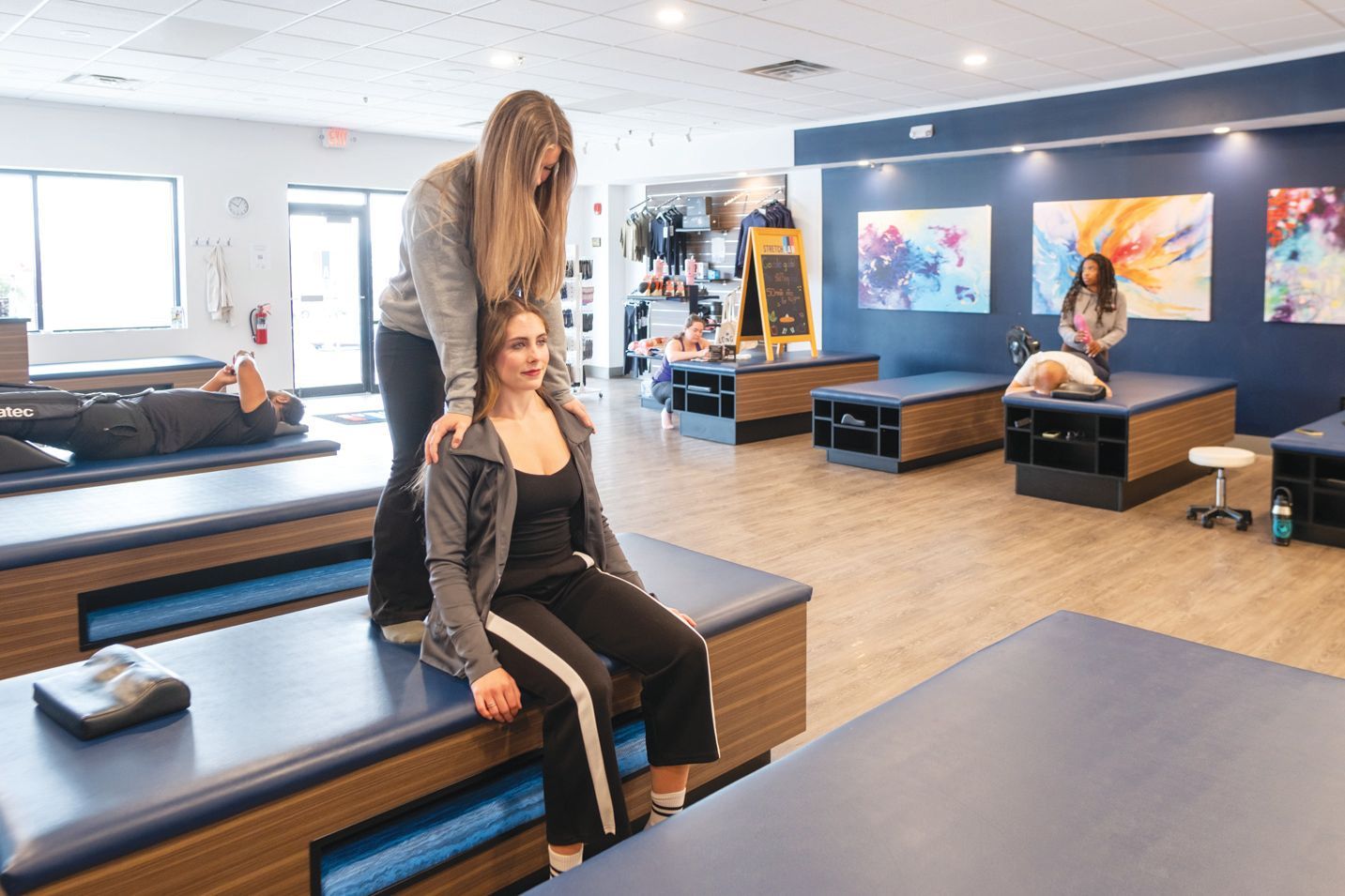 A therapist assists a client with a seated stretch on a padded treatment table in a clinic with abstract wall art.