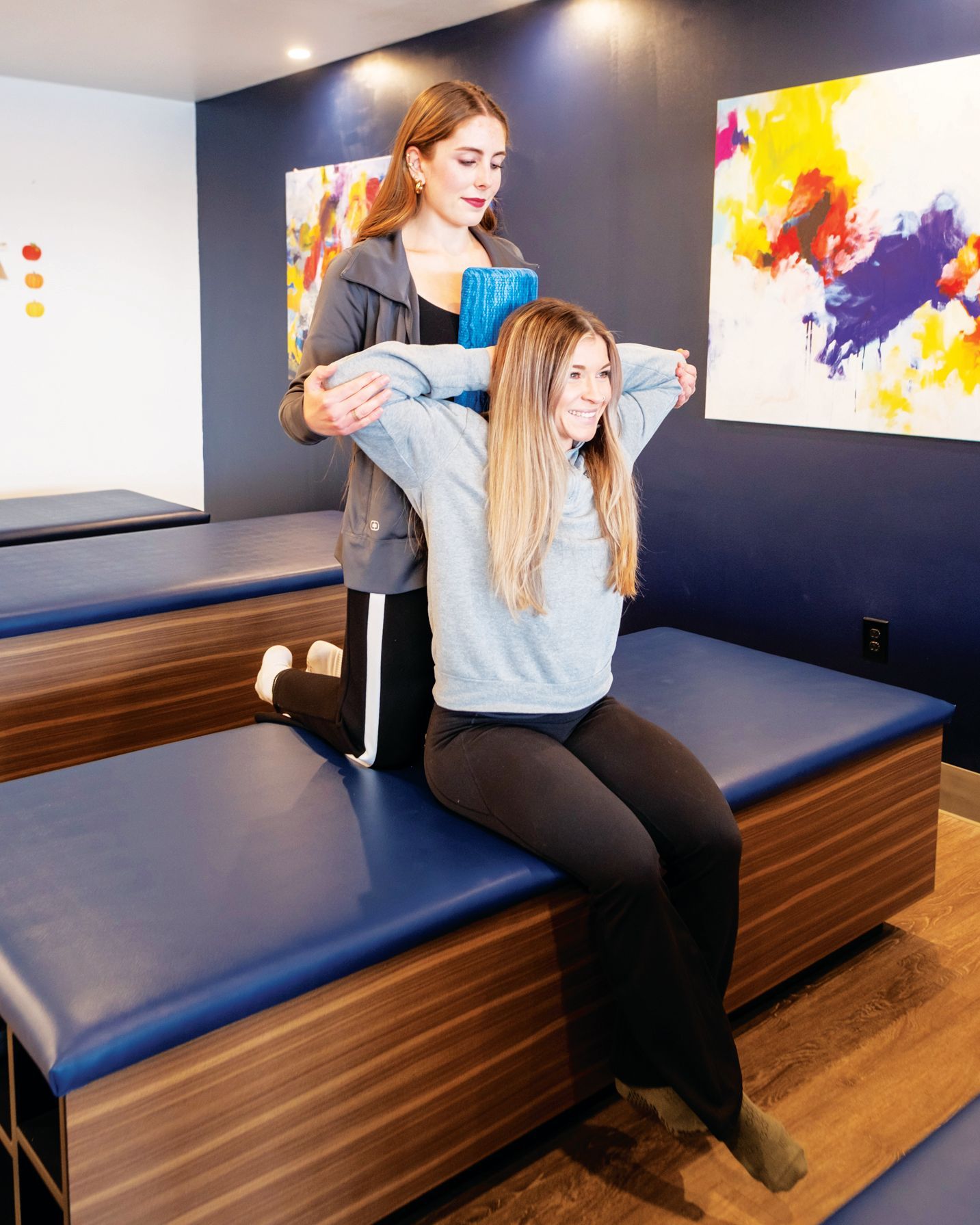 A physical therapist assists a patient performing a thoracic extension stretch using a blue foam roller on a treatment table.