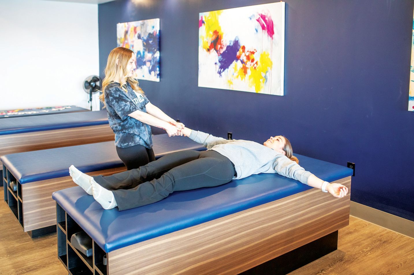 A practitioner holding a person's hands while they lie on a blue massage table in a room with abstract artwork.