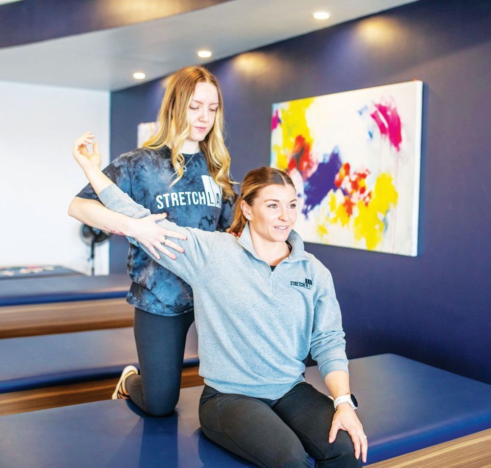 A practitioner assists a seated client with a shoulder stretch in a professional wellness studio.