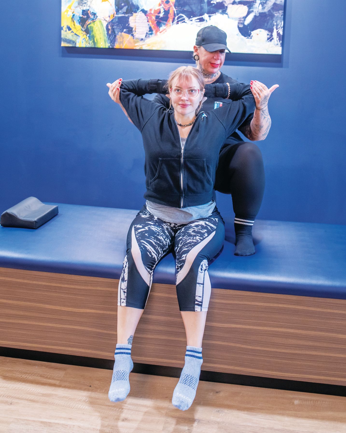 A physical therapist assists a patient in a seated upper-body stretch on a treatment table in a clinic.