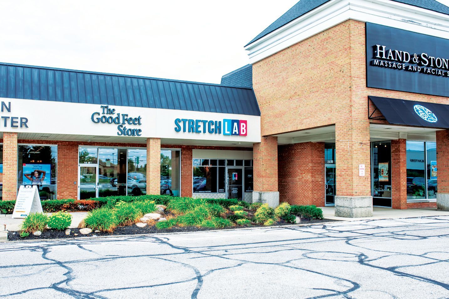 A brick shopping center strip with storefronts including The Good Feet Store, StretchLab, and Hand & Stone, under a roof.