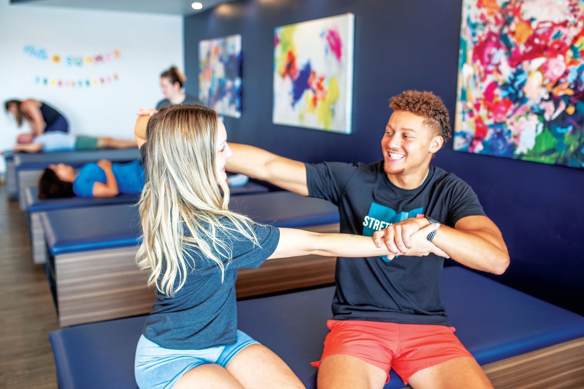 A professional assisted-stretching session where a practitioner guides a client's arm stretch in a brightly decorated studio.