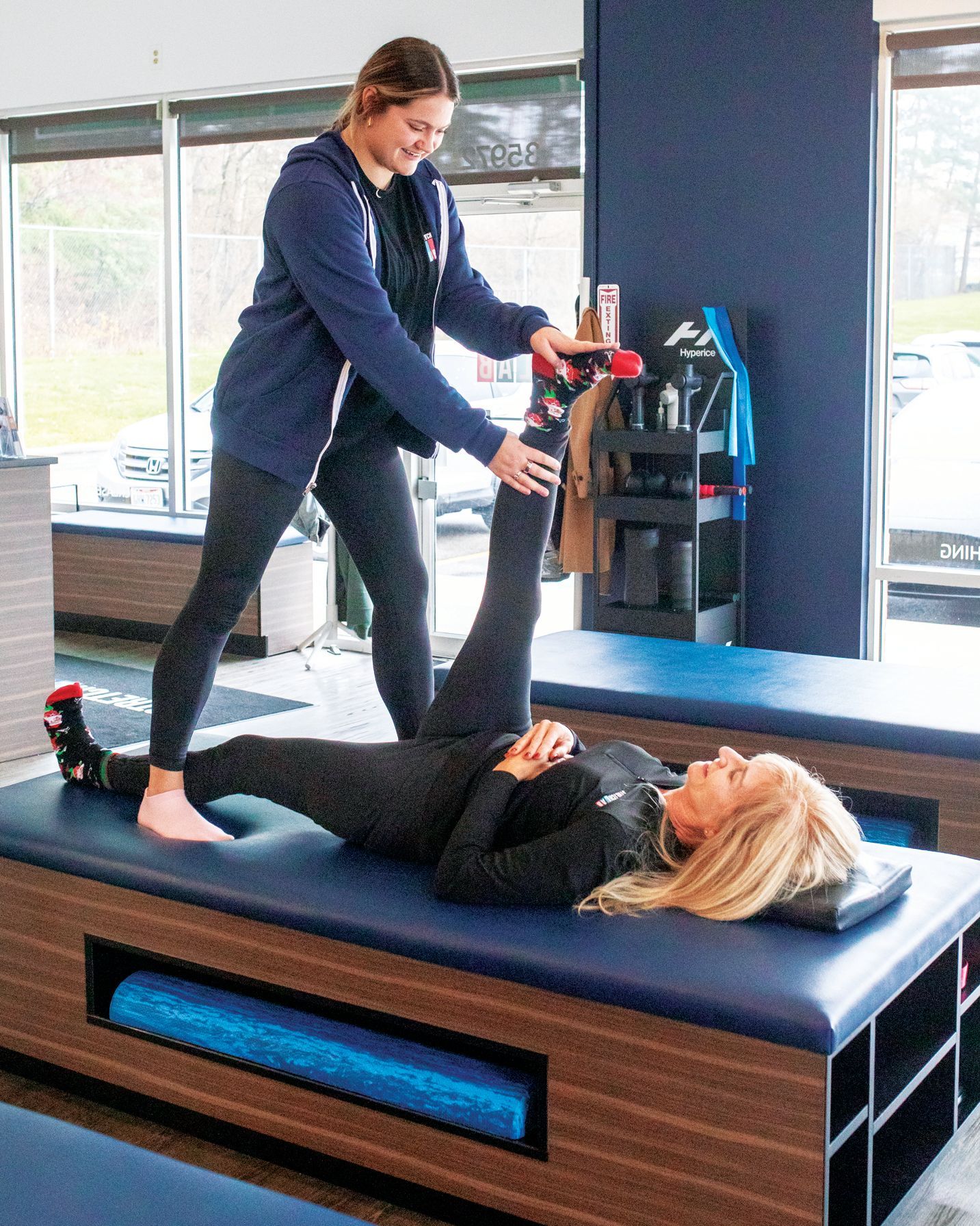 A professional assists a person lying on a physical therapy table by performing a supervised hamstring stretch.