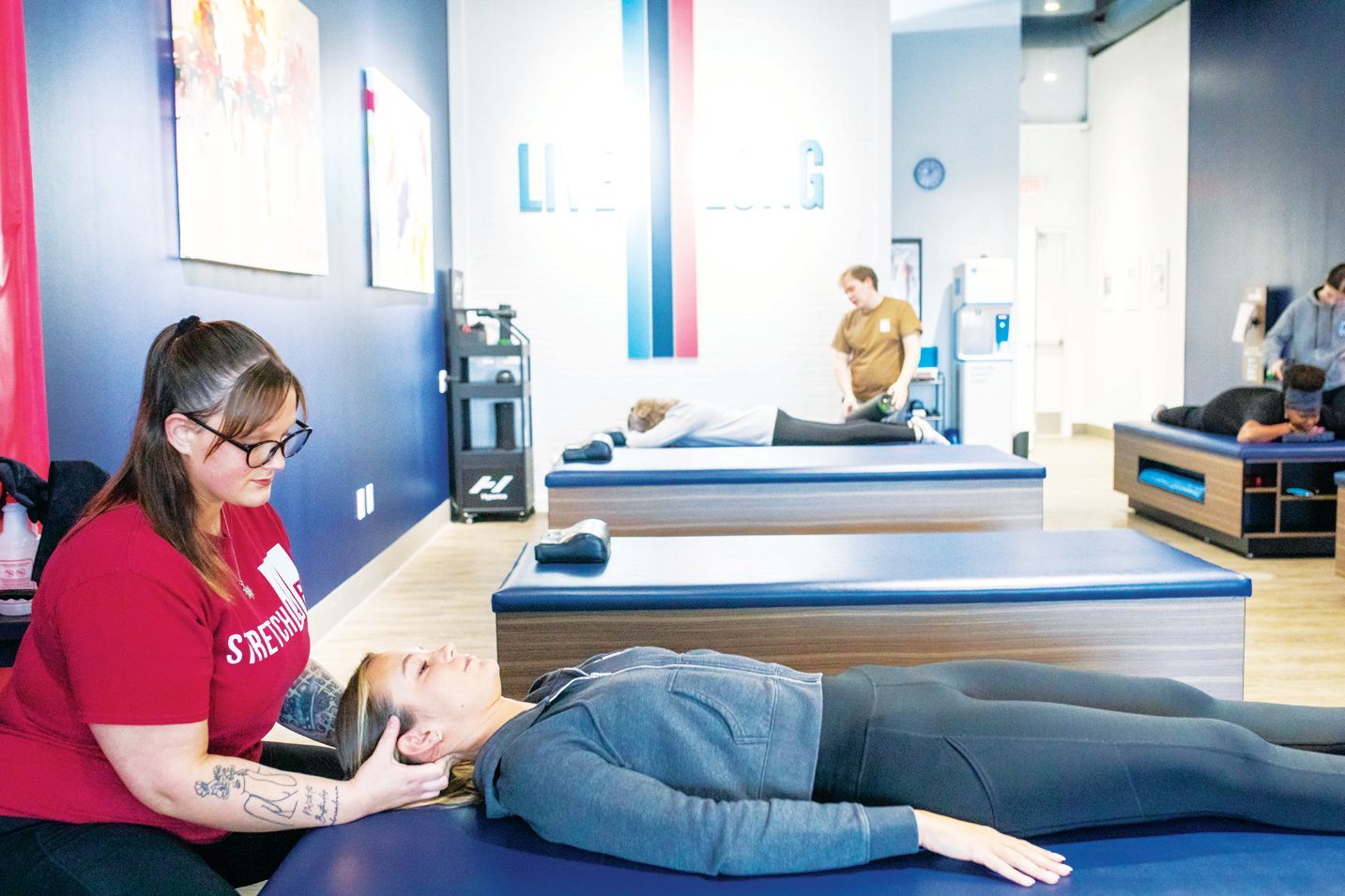 A practitioner in a red shirt performs assisted stretching on a client lying on a blue treatment table in a wellness studio.