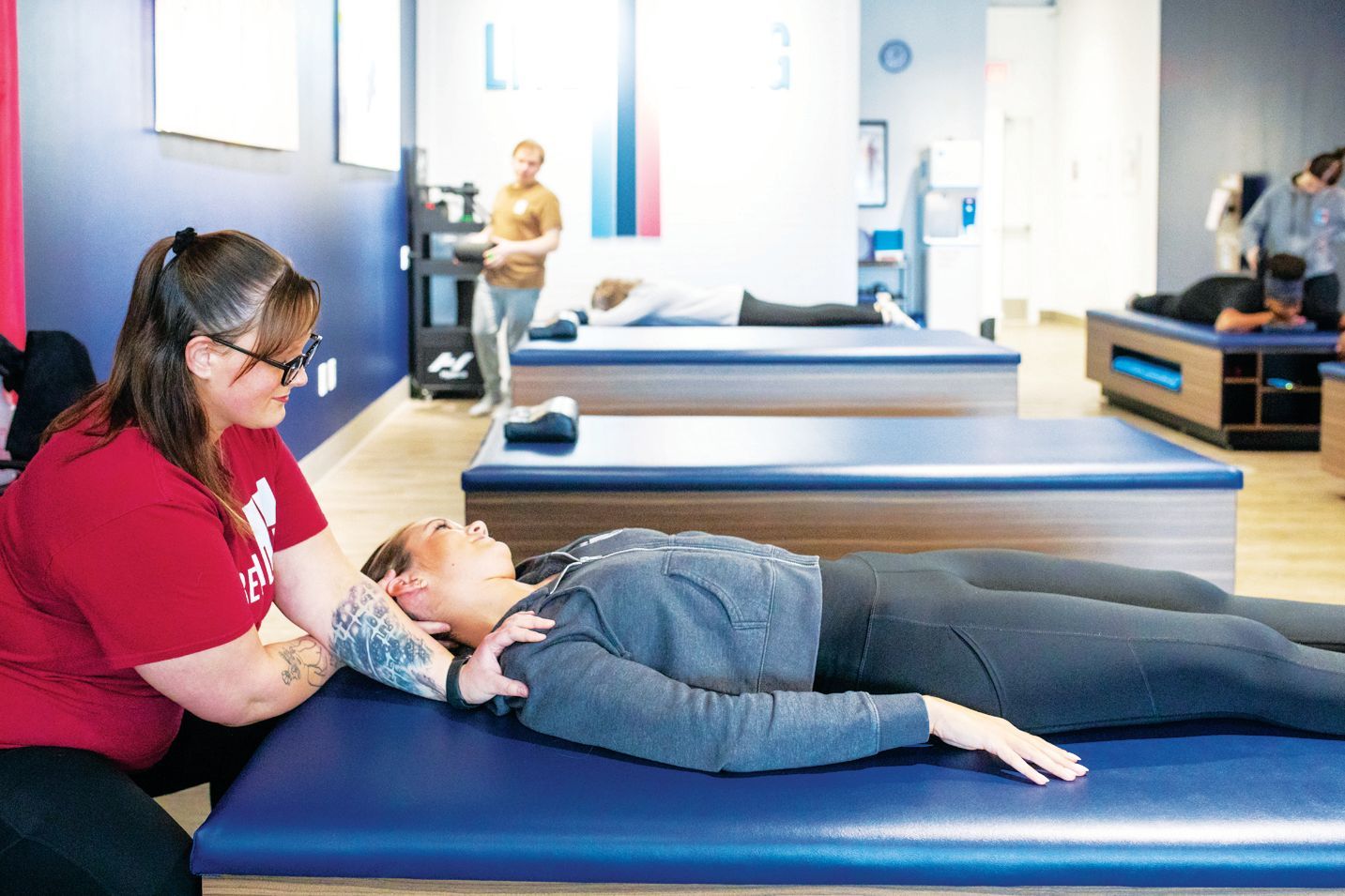 A therapist in a red shirt performs manual physical therapy on a patient lying on a blue treatment table in a clinic.