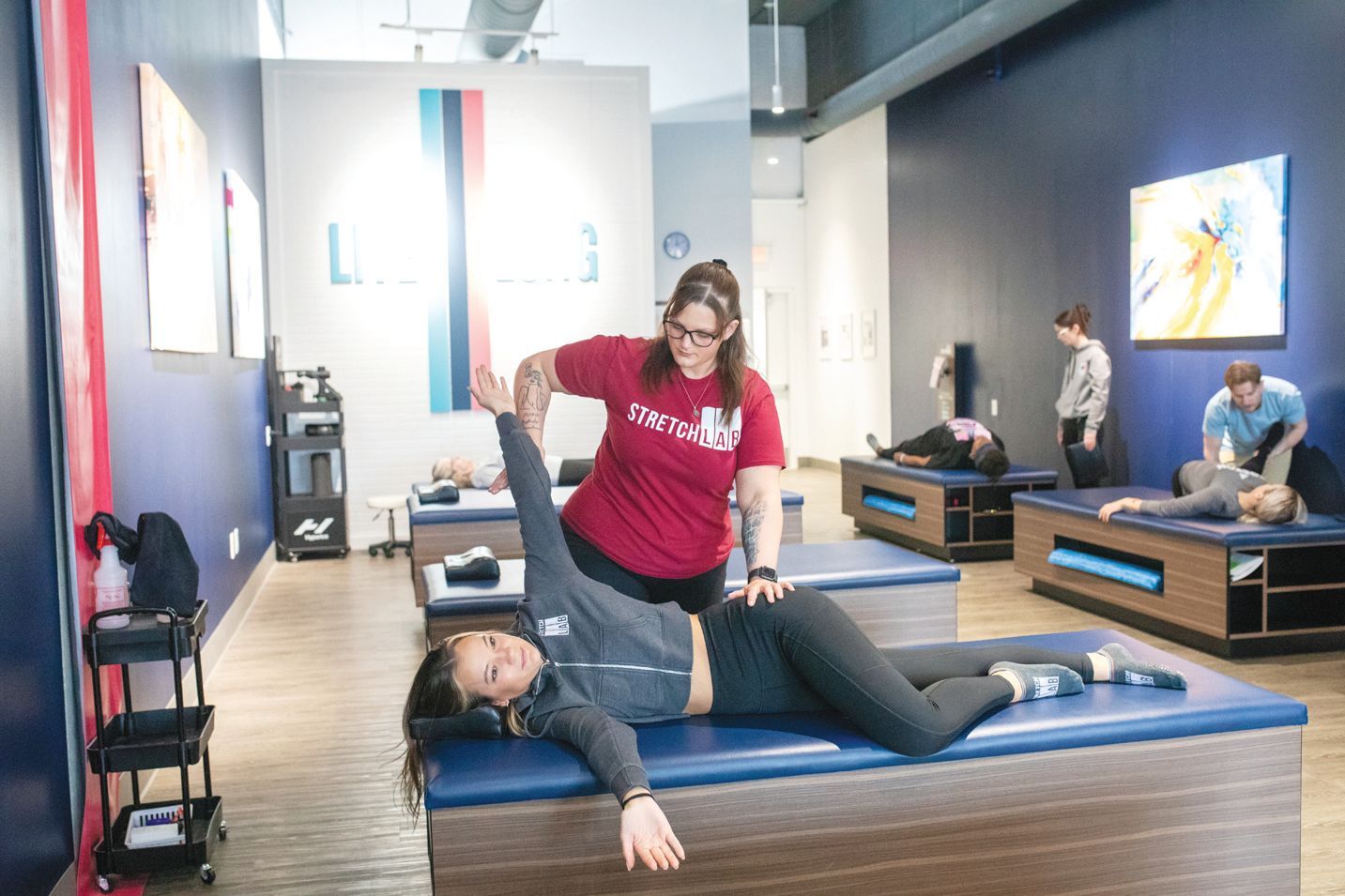 A practitioner in a red shirt guides a person through a stretching exercise on a blue massage table in a therapy studio.