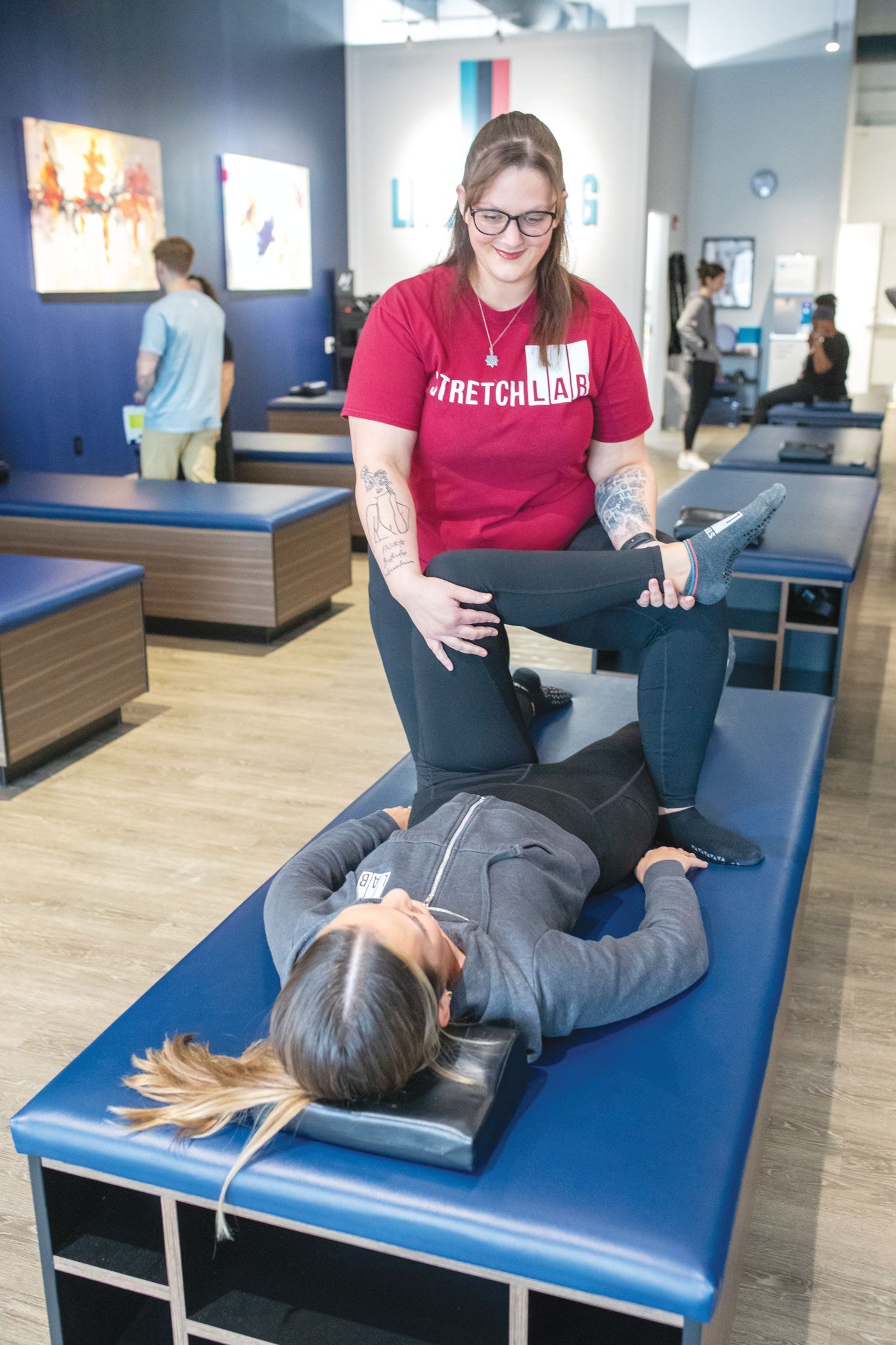 A practitioner in a red shirt performs a physical therapy stretch on a person lying on a blue table in a studio.