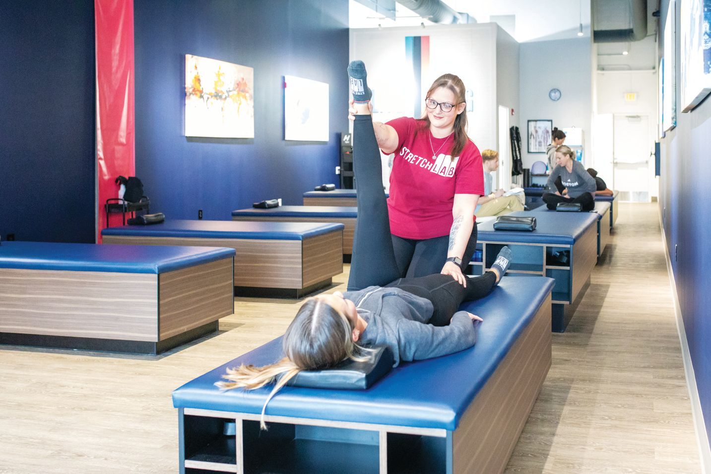 A professional stretches a person's leg on a table in a physical therapy clinic with blue walls and multiple tables.