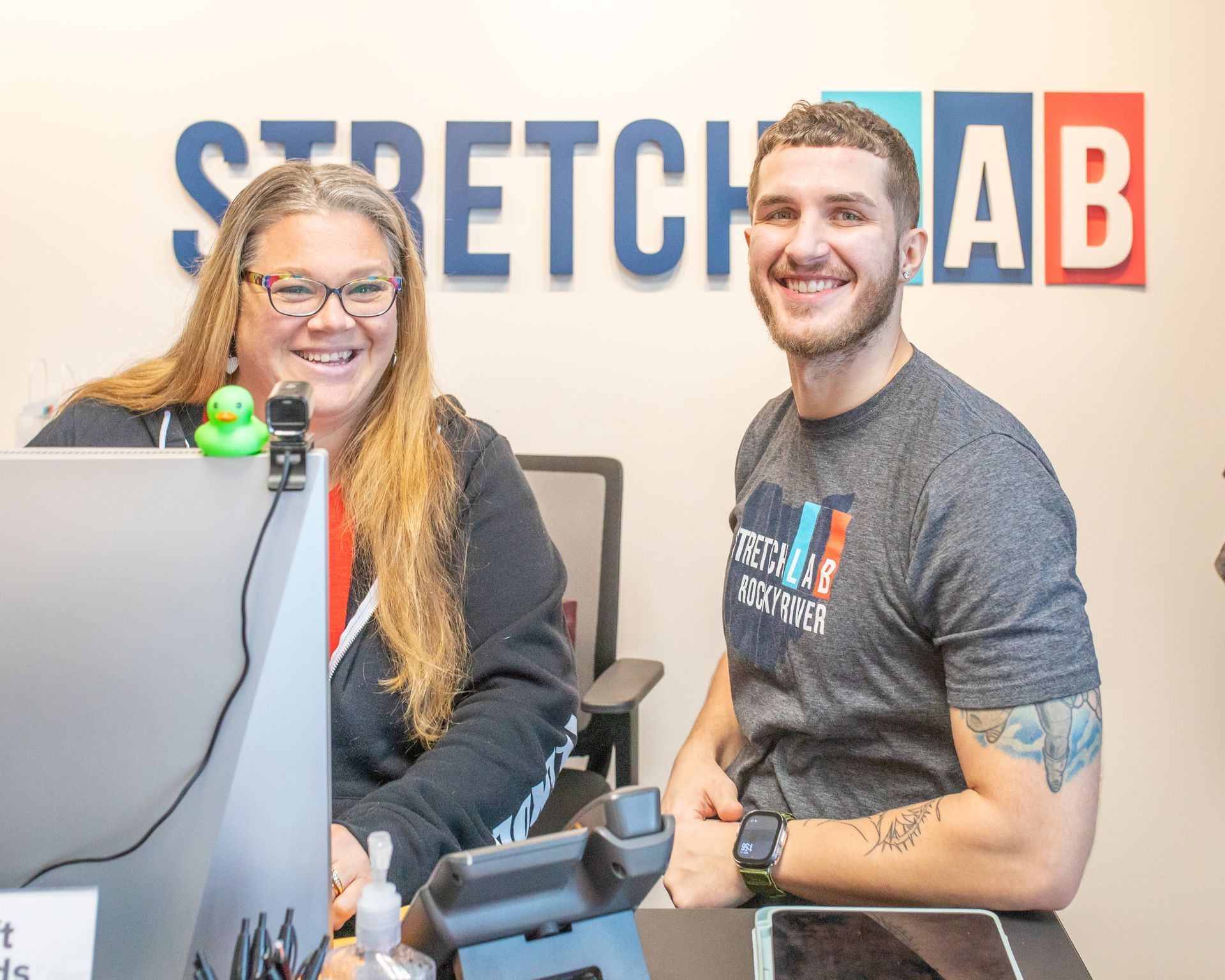 A person with long hair and glasses and a smiling person with a tattoo sitting together at a StretchLab front desk.
