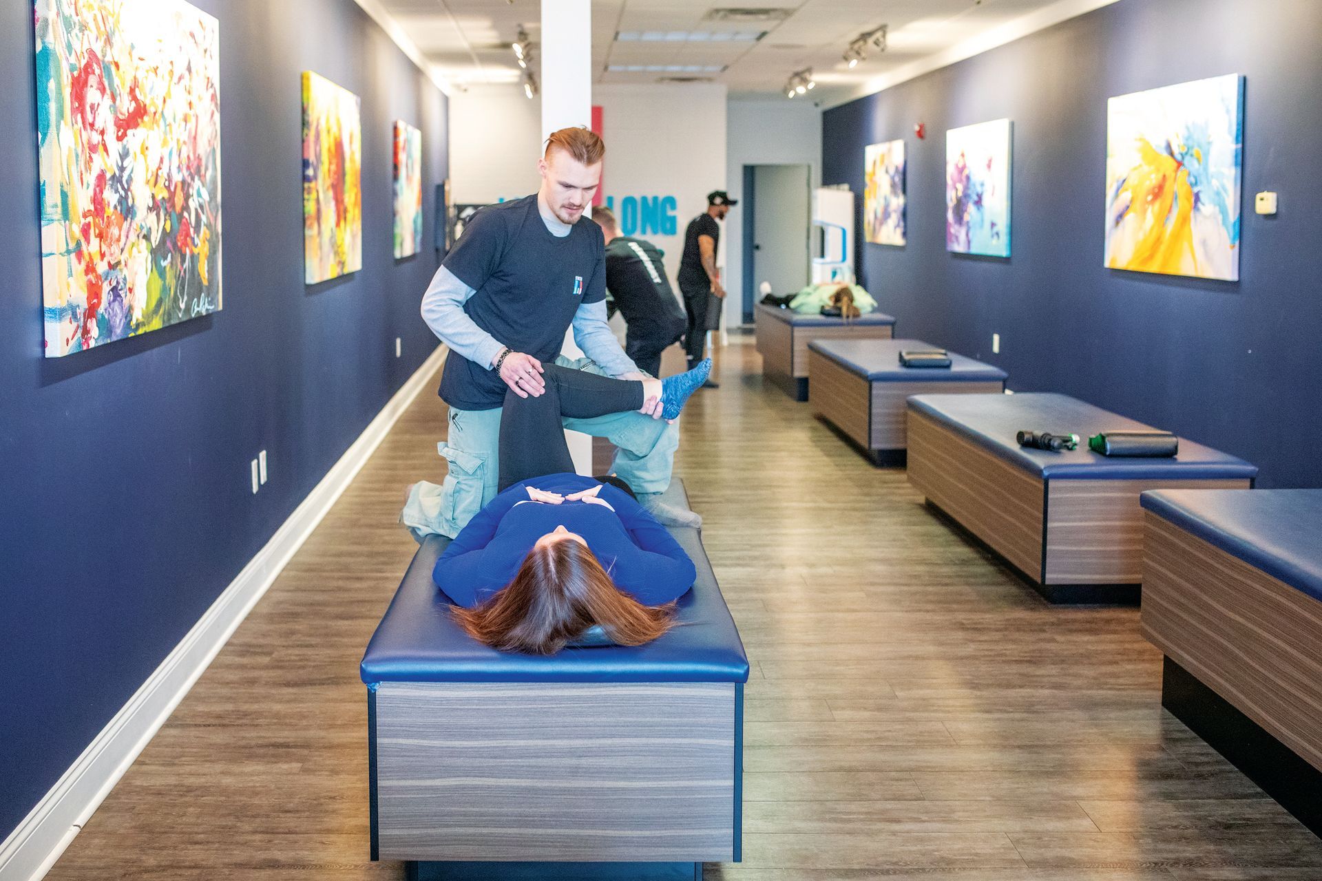 A professional helps a person on a therapy table in a studio with wood floors and art on navy blue walls.