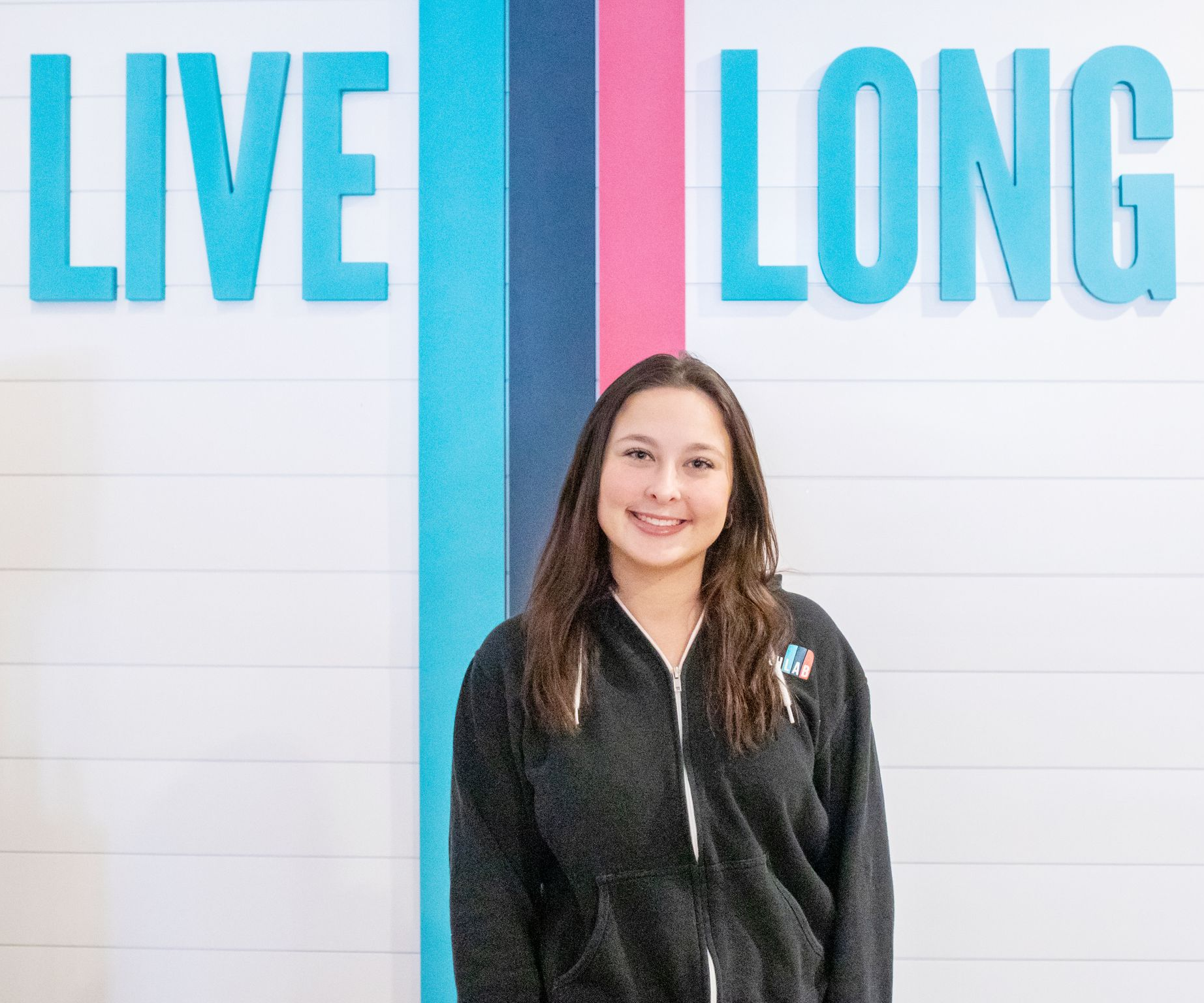 A person with long brown hair smiles in a dark zip-up hoodie against a white wall with the words 