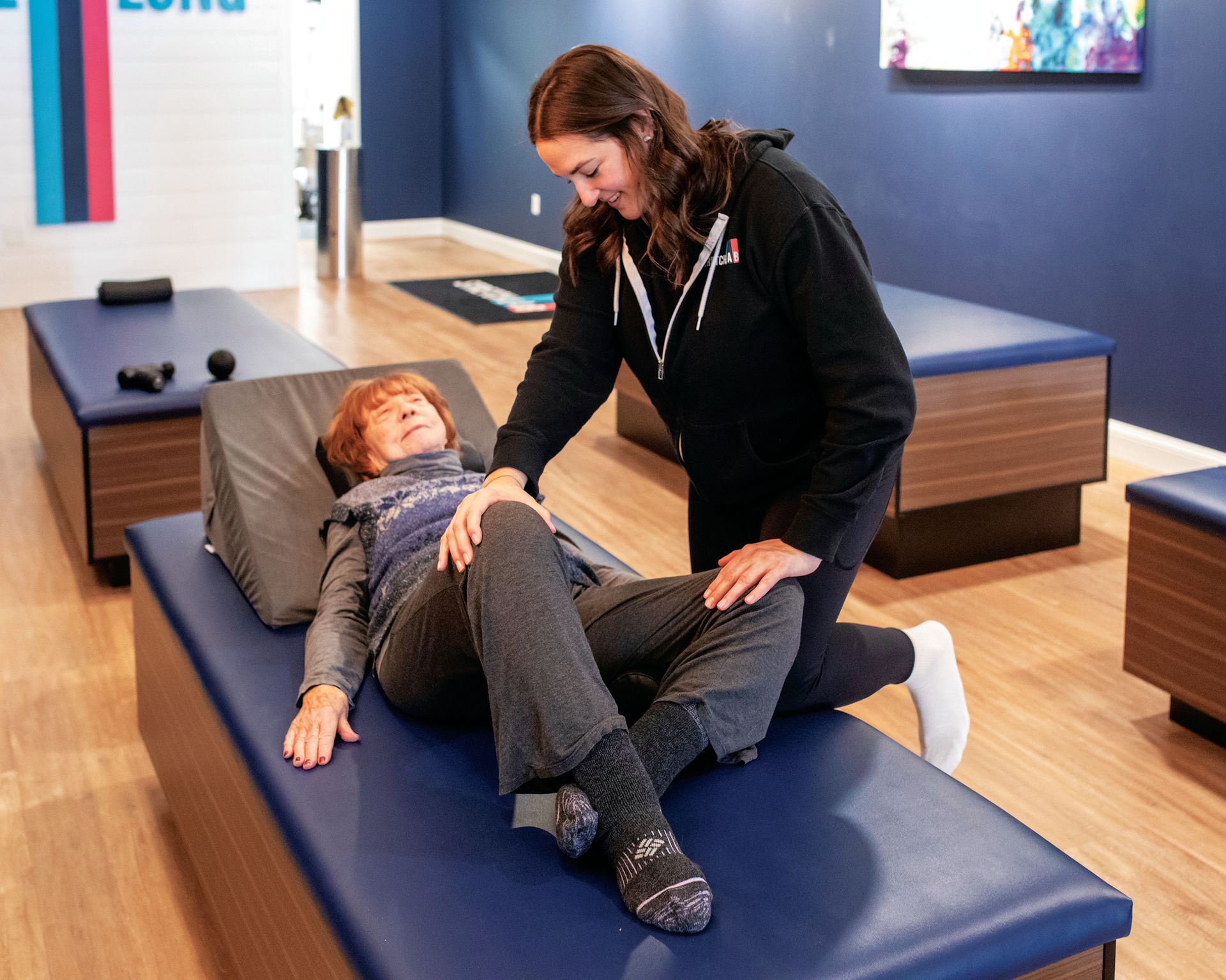 A physical therapist assists a client lying on a treatment table with a leg stretch in a clinic.