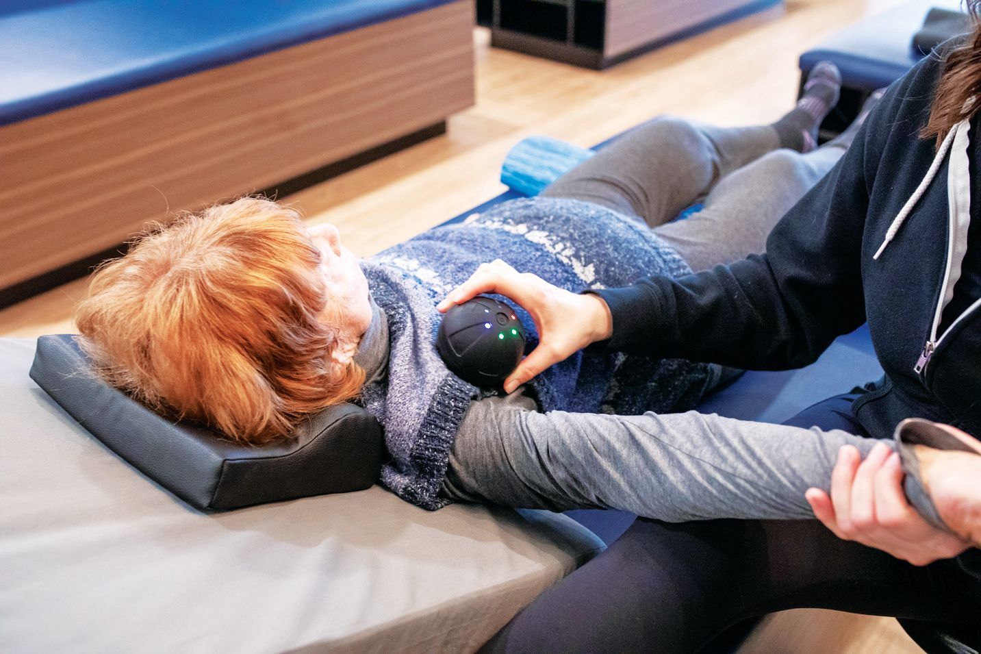 A physical therapist uses a vibrating massage ball on a patient’s shoulder while lying on a therapy table.