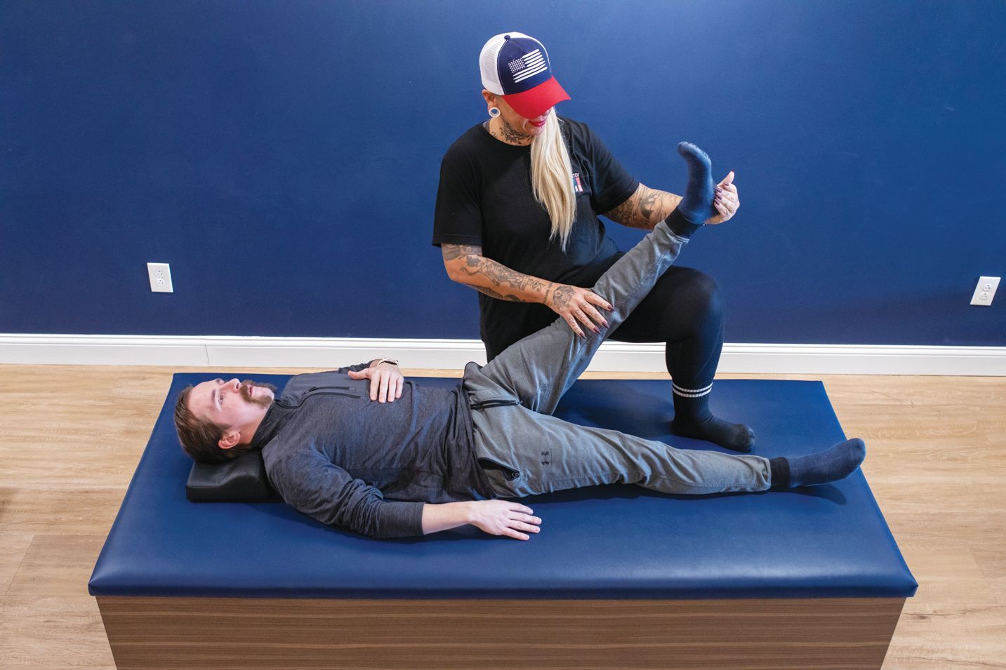 A person in a black shirt and cap performs a passive straight-leg stretch on a client lying on a treatment table.