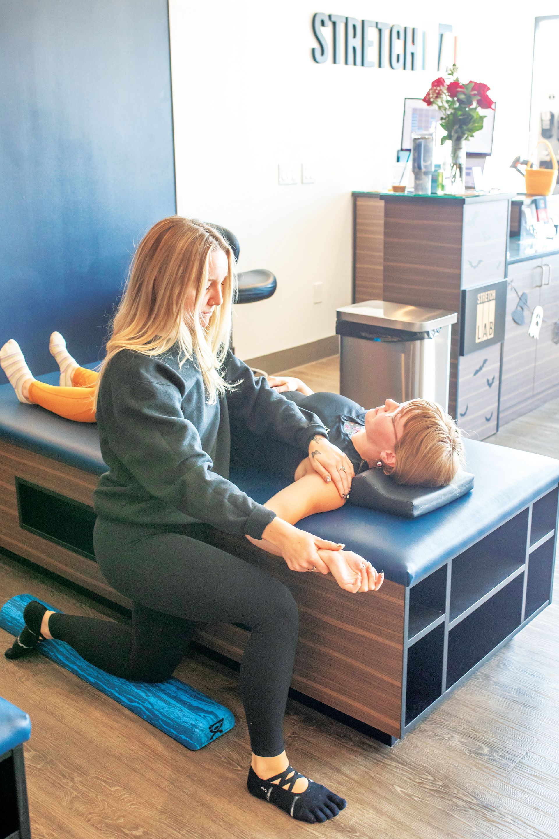 A practitioner performs assisted stretching on a client lying on a treatment table in a studio.