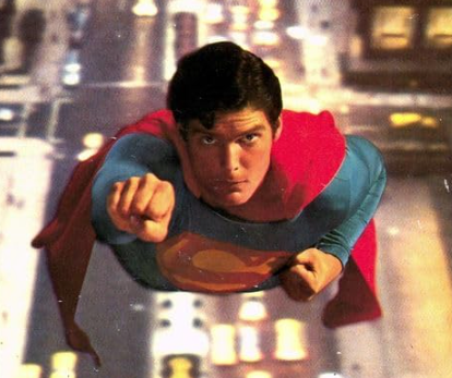 Superman flying forward over a blurry city street, wearing a blue suit with a red cape and chest emblem.