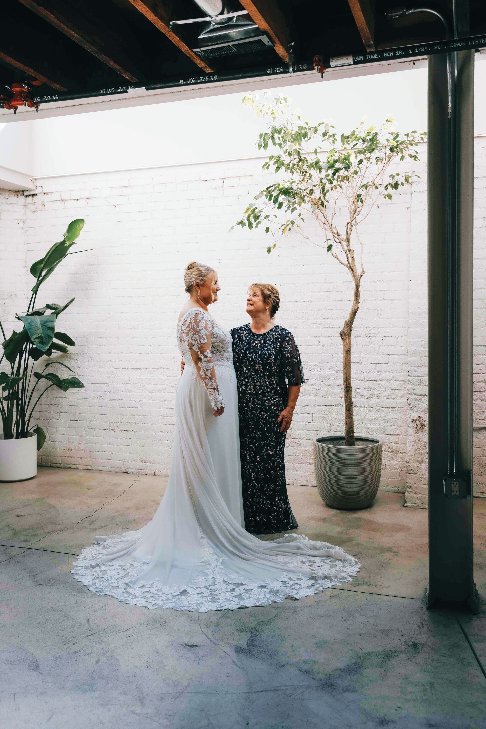 A bride in a long, white lace-sleeved dress stands facing an older woman in a sparkling, dark, long-sleeved gown.