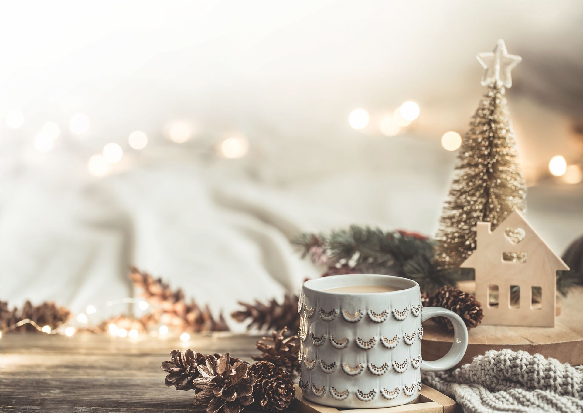 A cozy holiday scene featuring a patterned mug, pinecones, a small decorative house, and a miniature tree with soft lights.