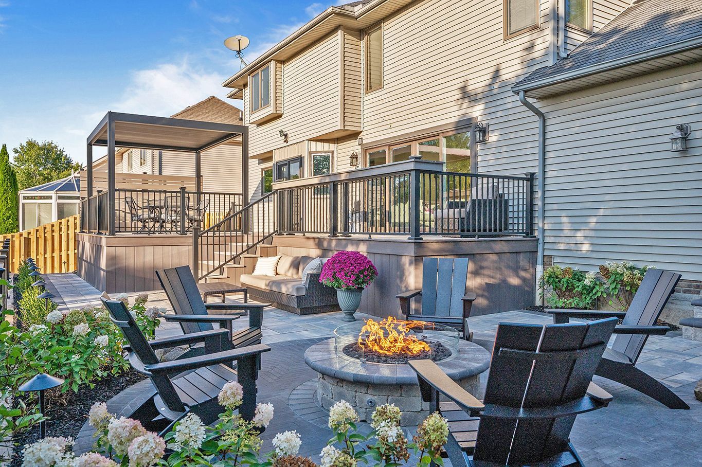 A backyard patio features a circular stone fire pit surrounded by Adirondack chairs, leading to a multi-level deck.