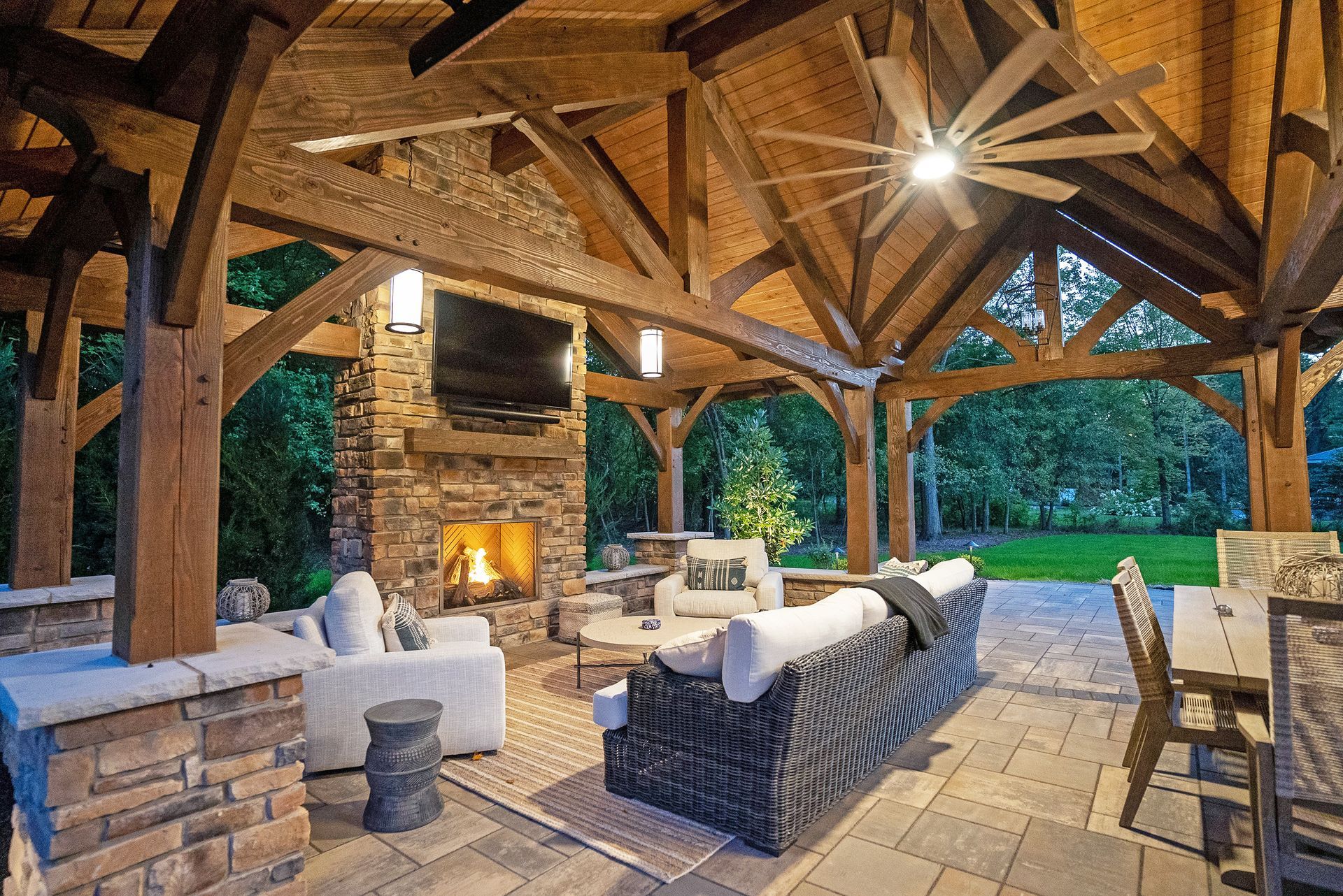 A luxury outdoor covered patio with a stone fireplace, television, rustic wooden beams, and lounge seating.