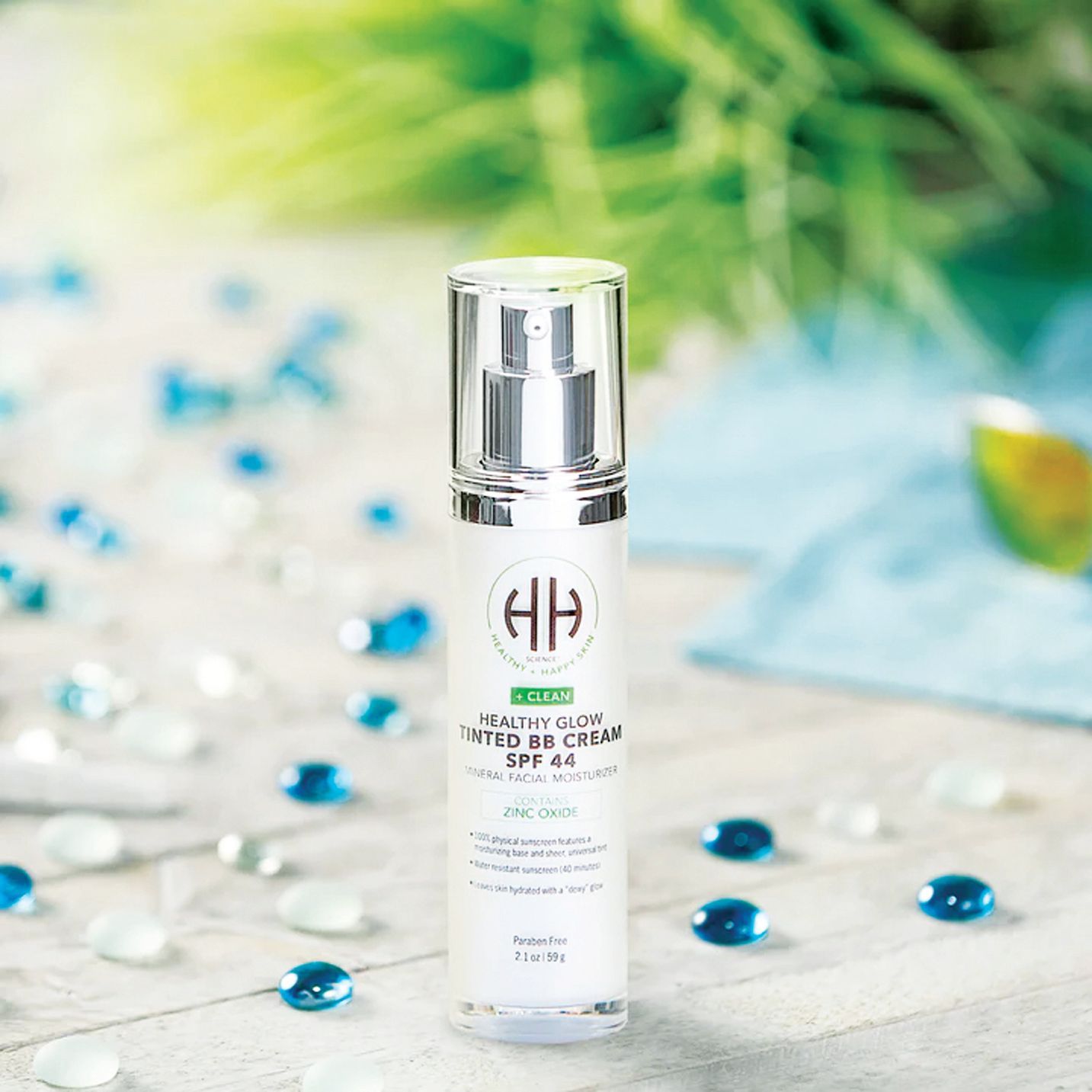 A bottle of HH Healthy Radiance Tinted BB Cream SPF 44 standing on a white wooden surface with scattered blue glass beads.