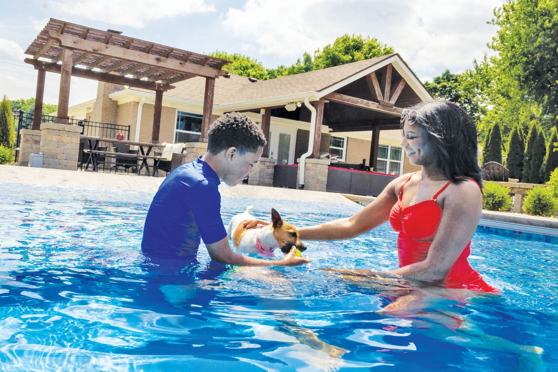 Two people play with a small dog in a backyard swimming pool in front of a pergola and patio area.