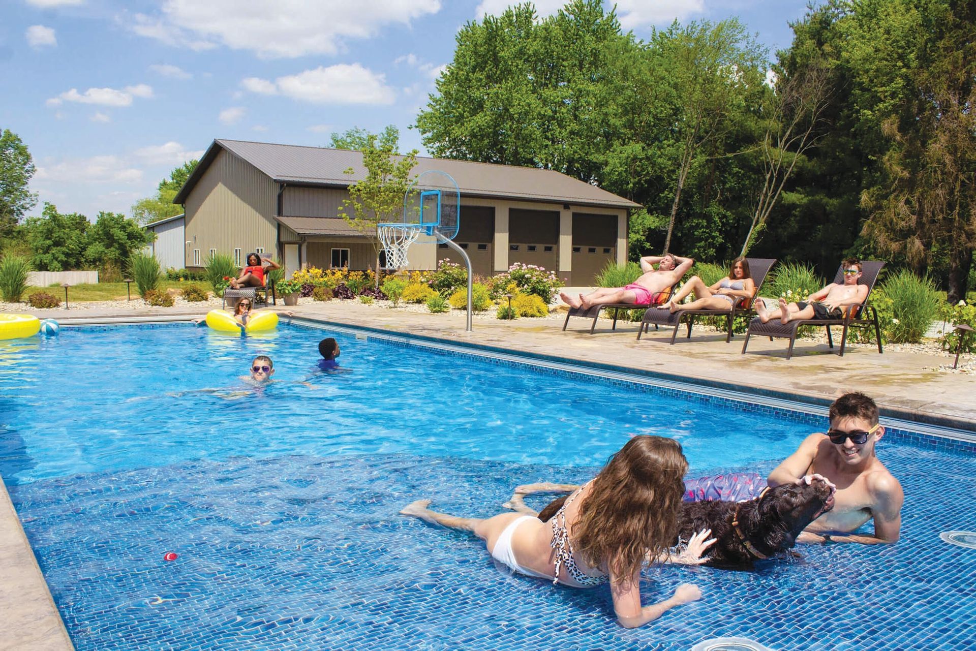 People relax in a sunny outdoor pool with a dog, while others lounge on chairs in front of a building on a summer day.