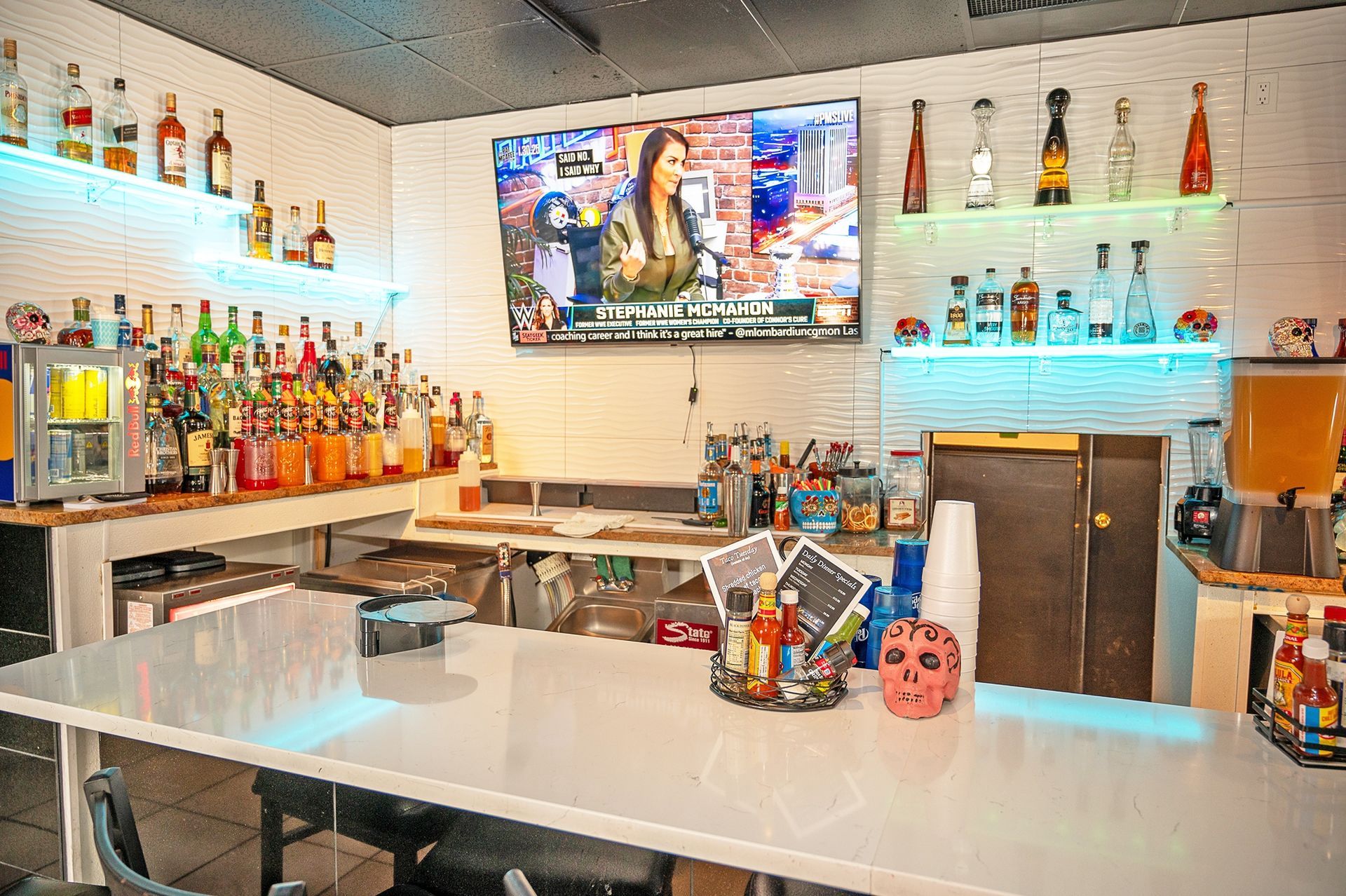 A white bar counter with a pink skull decoration, stocked shelves with bottles, and a large television on the wall.