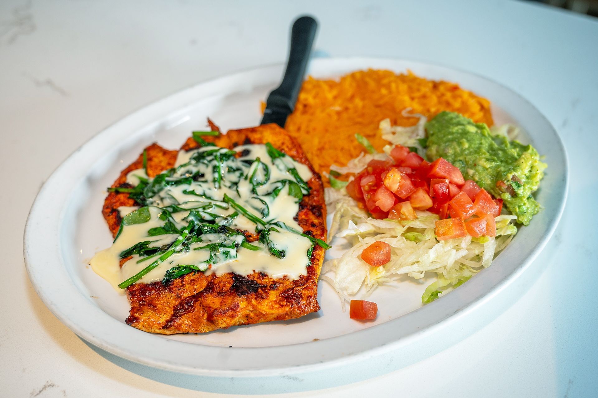 A platter featuring grilled chicken topped with spinach and cheese, served with rice, shredded lettuce, tomato, and dip.
