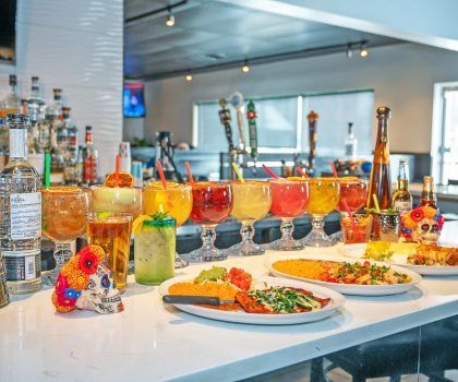 A bar counter features several colorful cocktails in large glasses, plates of Mexican food, and a decorative sugar skull.
