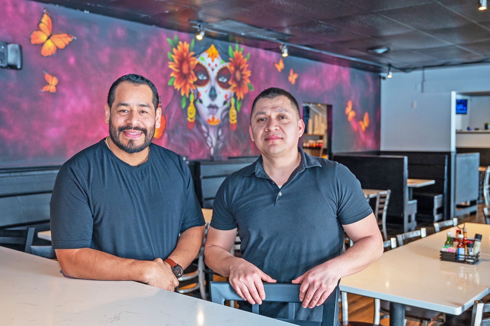 Two people stand in a restaurant with a mural of a sugar skull on the wall behind them.