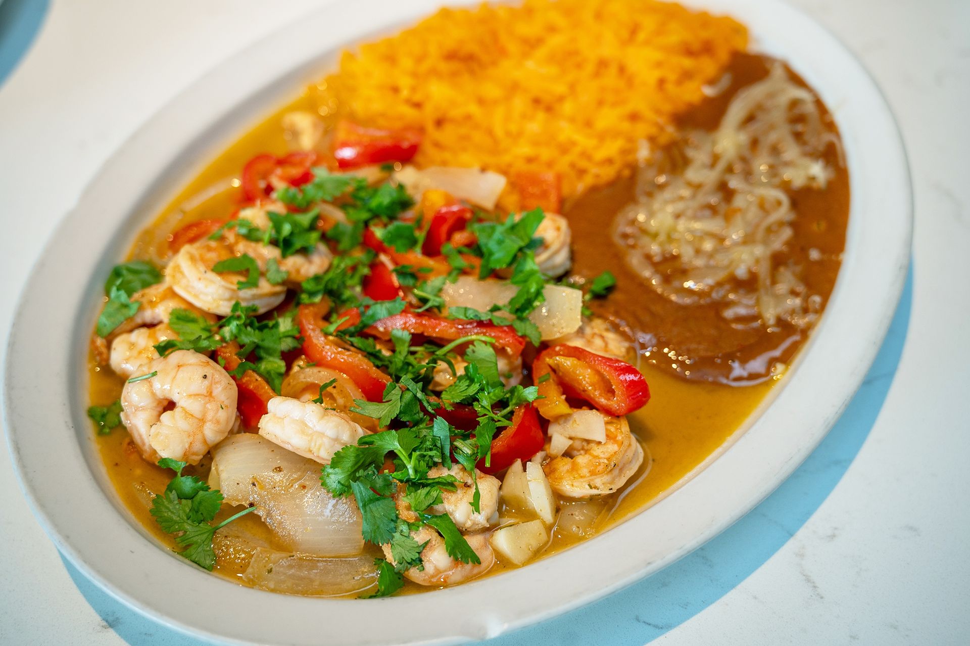 A shrimp fajita dish with red peppers, onions, cilantro, Mexican rice, and refried beans on an oval white plate.
