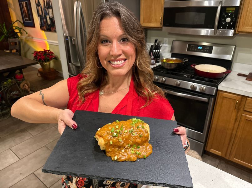 A smiling person in a red top holds a slate board featuring a food dish covered in sauce and herbs in a home kitchen.