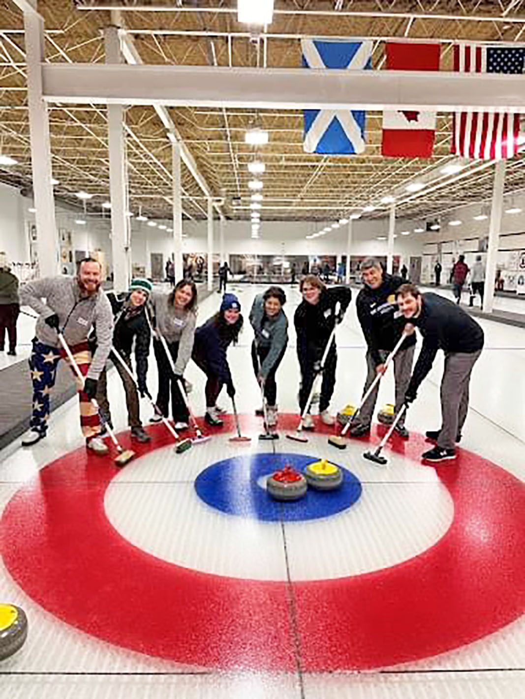 A group of people standing on a curling rink, holding brooms and posing behind curling stones on the house target.
