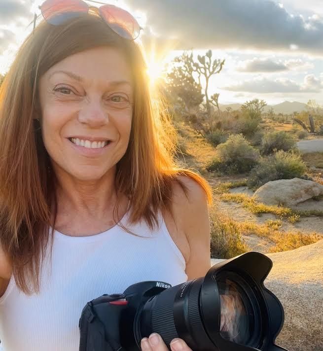 A person holding a camera smiles while standing in a desert landscape during a sunset with sun rays in the background.