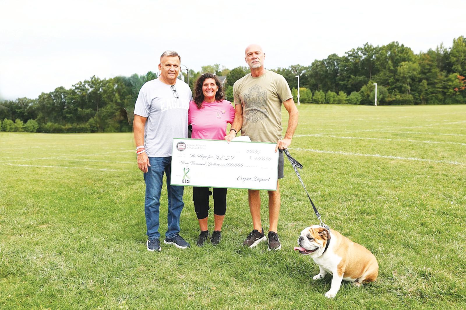 Three people holding a large donation check stand in a grassy field next to an English Bulldog.