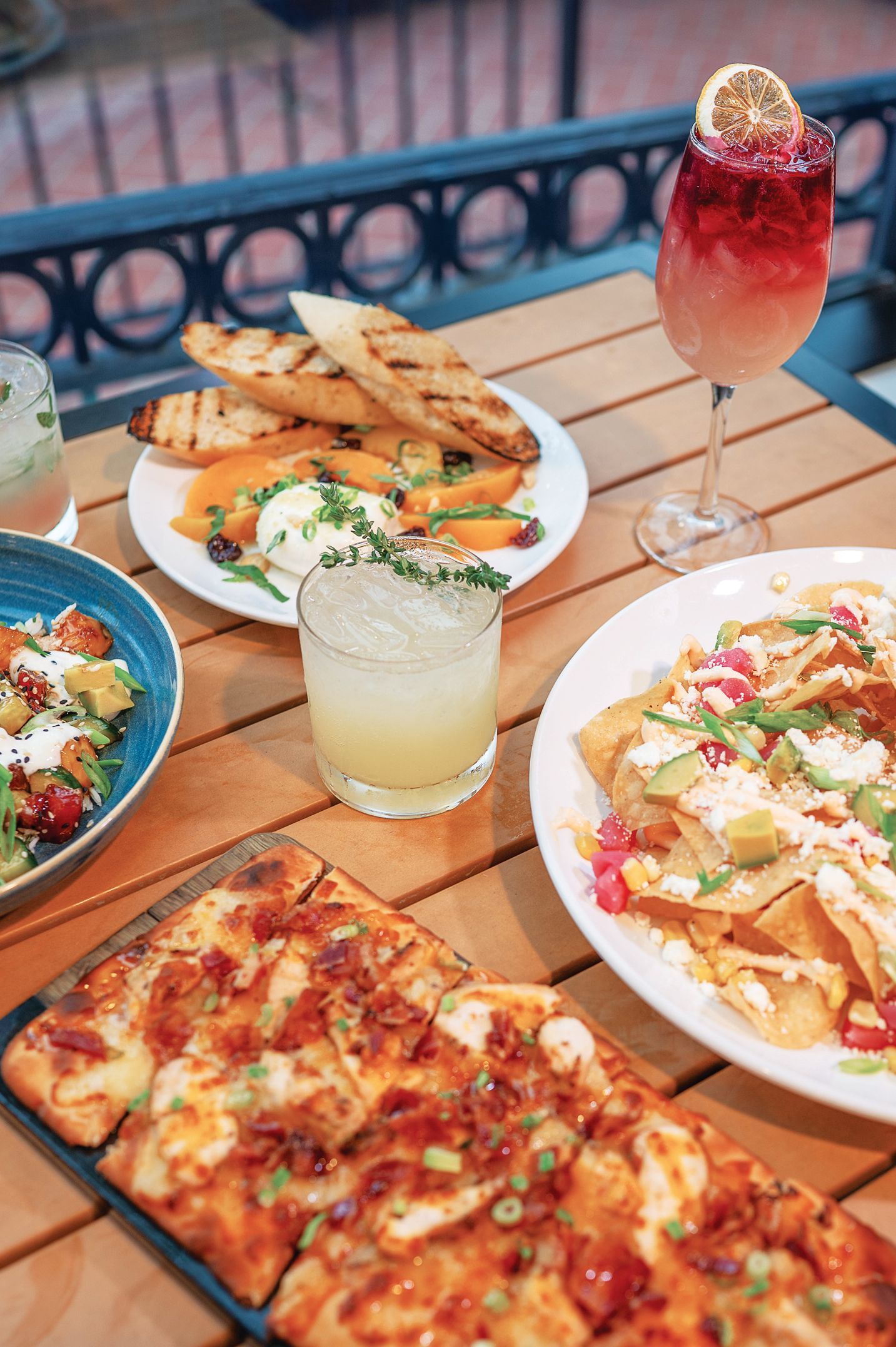 A wooden table filled with plates of flatbread, nachos, and grilled bread, alongside two colorful cocktails.