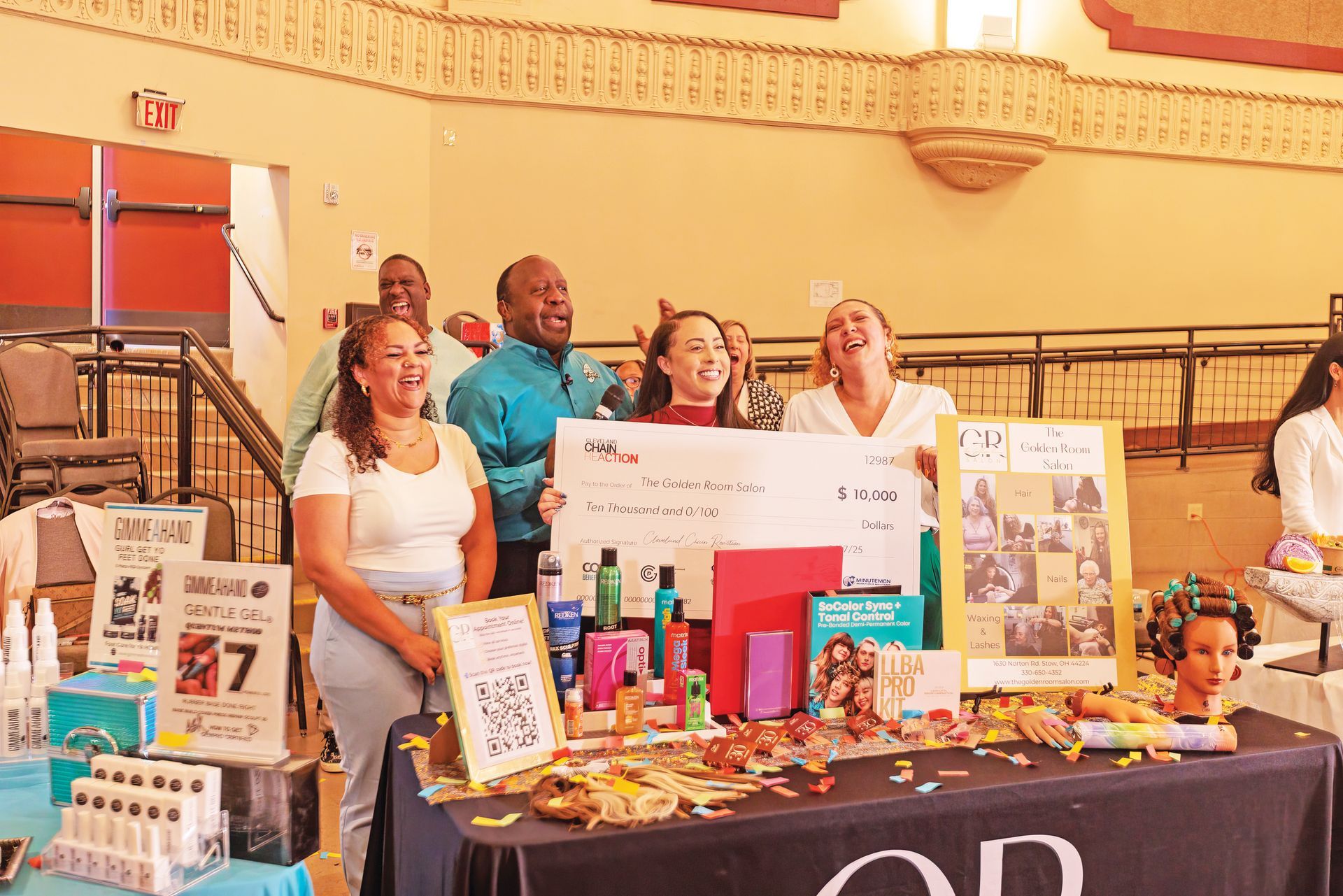 Four people celebrate behind a display table at an event, holding a large check with various beauty products on the table.