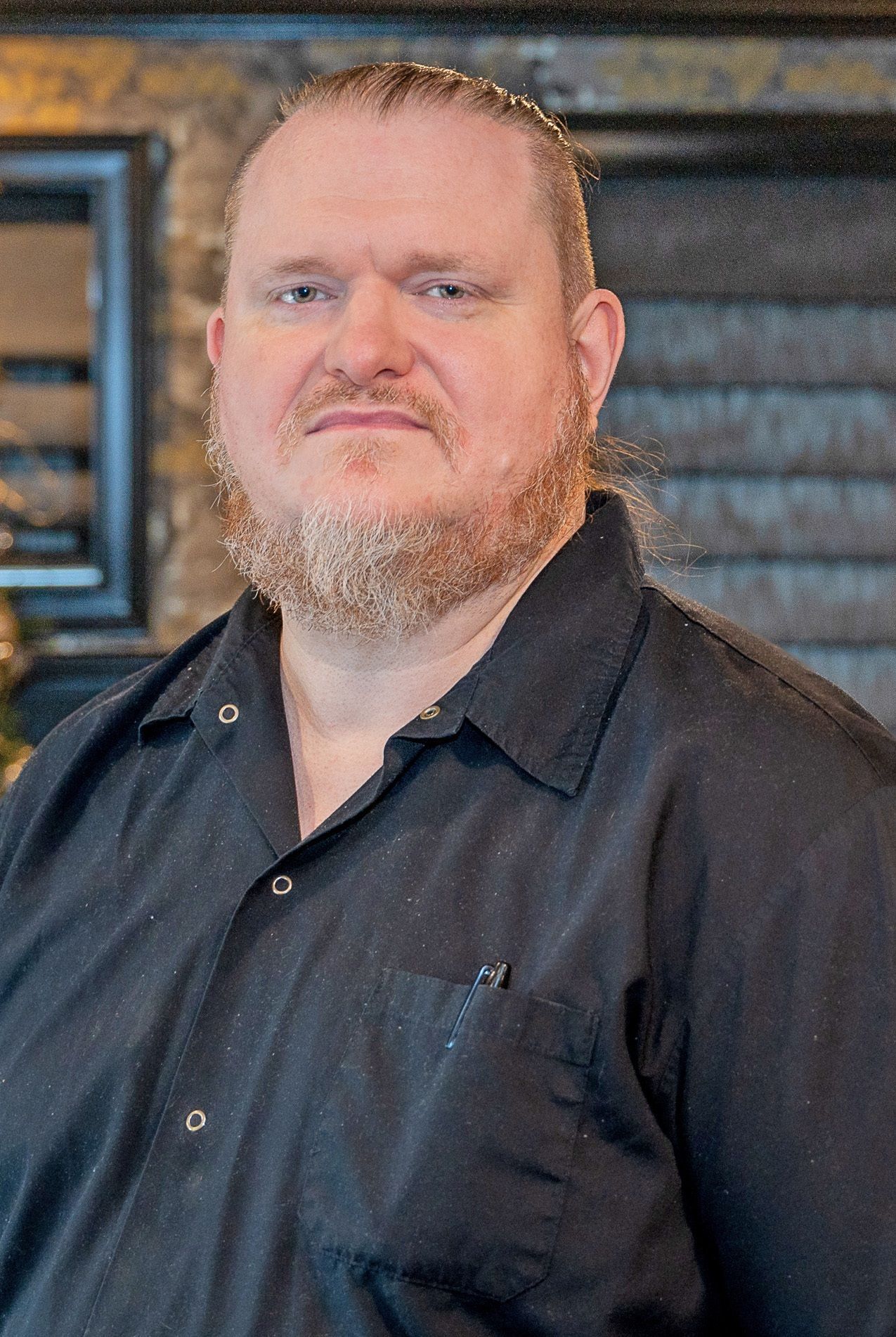 A person with a beard, wearing a black collared shirt, poses for a portrait against a textured background.