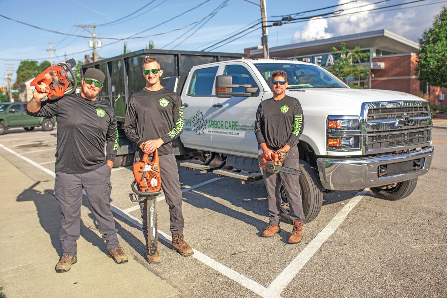 Three tree service workers in uniform with chainsaws stand in a parking lot next to their white dump truck.