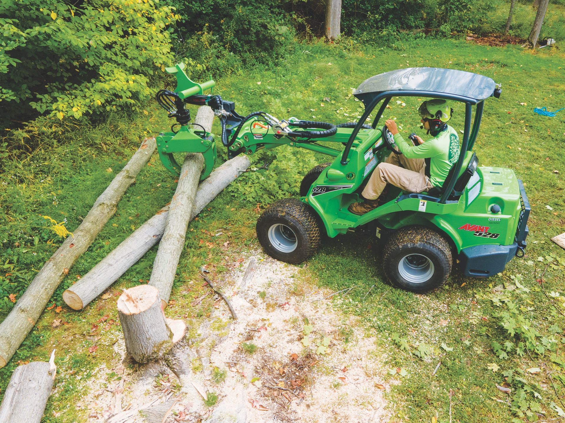 A person in a helmet operates a small, green utility loader with a grapple attachment to lift a log in a grassy area.