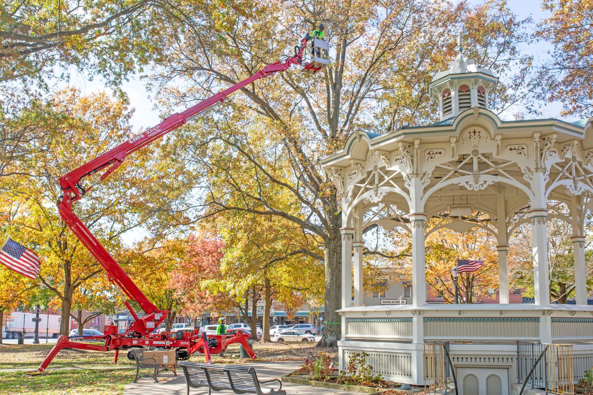 A red articulating boom lift reaching into the trees next to a white wooden gazebo in an autumn park.