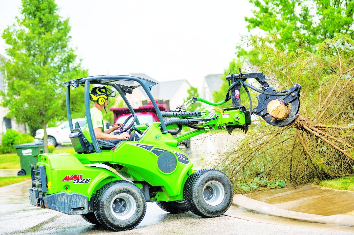 A bright green compact loader with a grapple attachment moves a fallen tree branch on a suburban street.