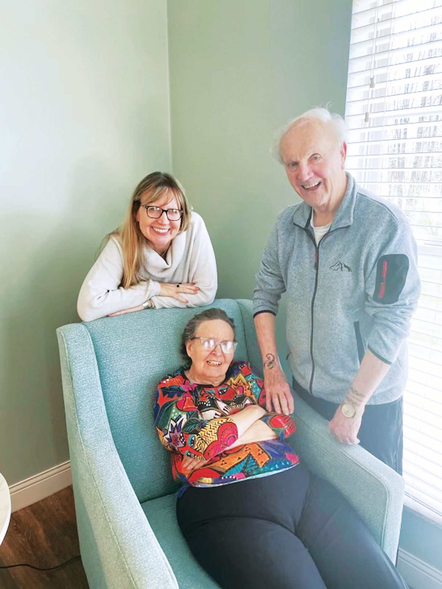 Three people smiling together: one seated in a teal armchair and two standing behind it in a room with light green walls.