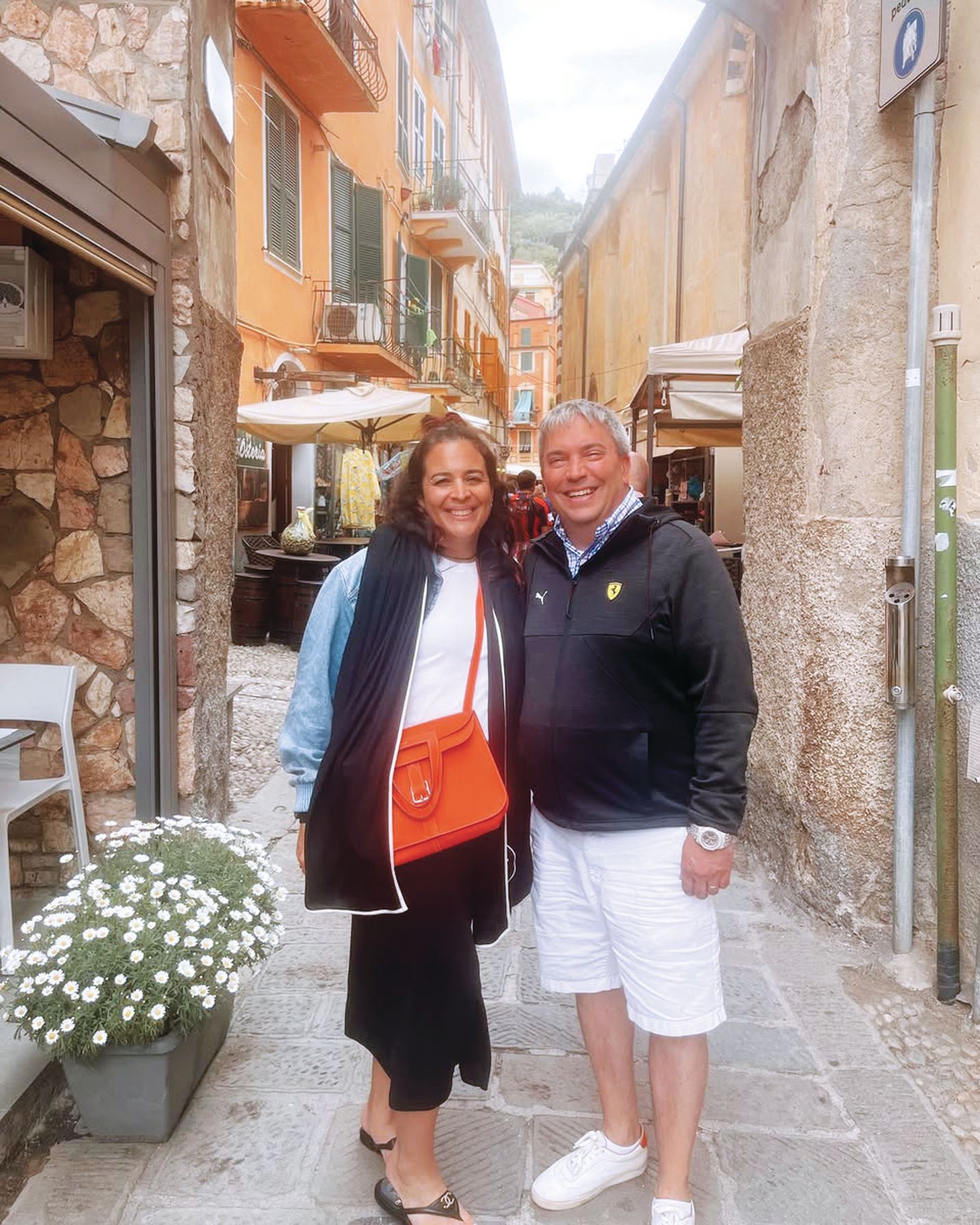 Two people stand smiling together on a narrow, stone-paved cobblestone street lined with colorful historic buildings.